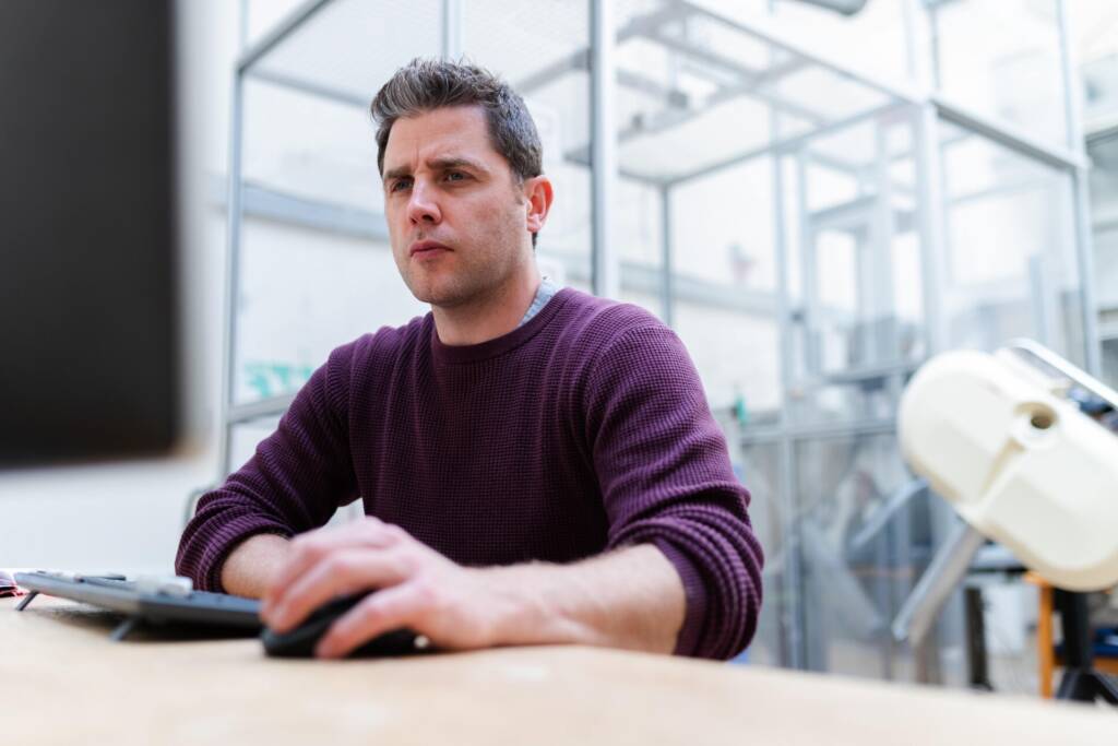 Professionele man in een donkerpaarse trui die aandachtig naar een computerscherm kijkt terwijl hij aan een bureau in een kantoor met glazen wanden werkt.